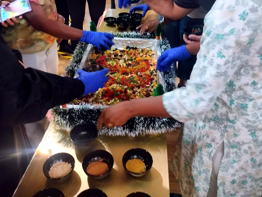 Gloved hands mixing dried fruit in a tinsel-trimmed tray, with bowls of spices in front